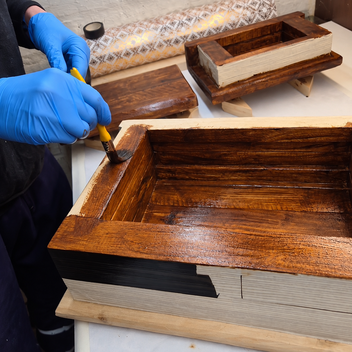Gloved hands brushing wood stain onto the rim of an Embergarth wooden book storage box, with its open interior and grooved page edges visible.