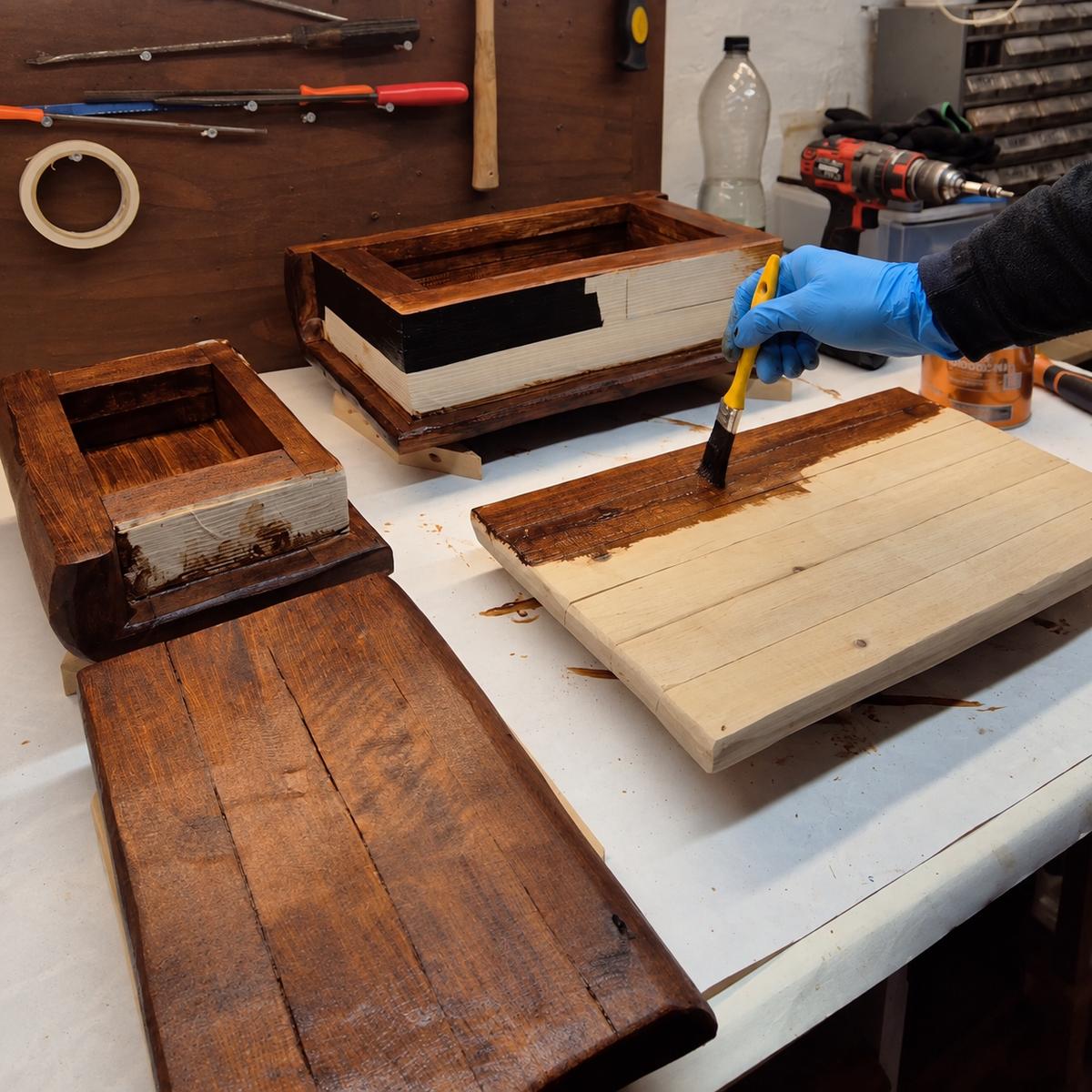 A gloved hand staining a wooden book cover beside open Embergarth book storage boxes and covers on the workshop bench.